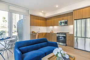 A modern kitchen with wooden cabinets, stainless steel appliances, and a blue sofa in a living area. - Sleeps 64 | Rooftop Pool + Spa + Fitness Studio + Bay Views + Walk to Dining (San Diego)