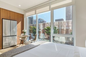 A modern bedroom with a large bed, a built-in refrigerator, a small dining table, and a view of the city through large windows. - Sleeps 64 | Rooftop Pool + Spa + Fitness Studio + Bay Views + Walk to Dining (San Diego)