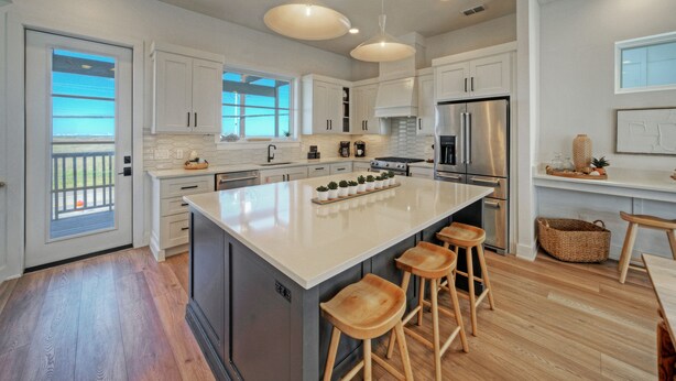A modern kitchen with white cabinets, a large island with stools, stainless steel appliances, and a view to the outside. - 4 Coastal Homes • Pools + Golf Cart Boardwalk Access | Dorado Dunes Collective (Port Aransas)