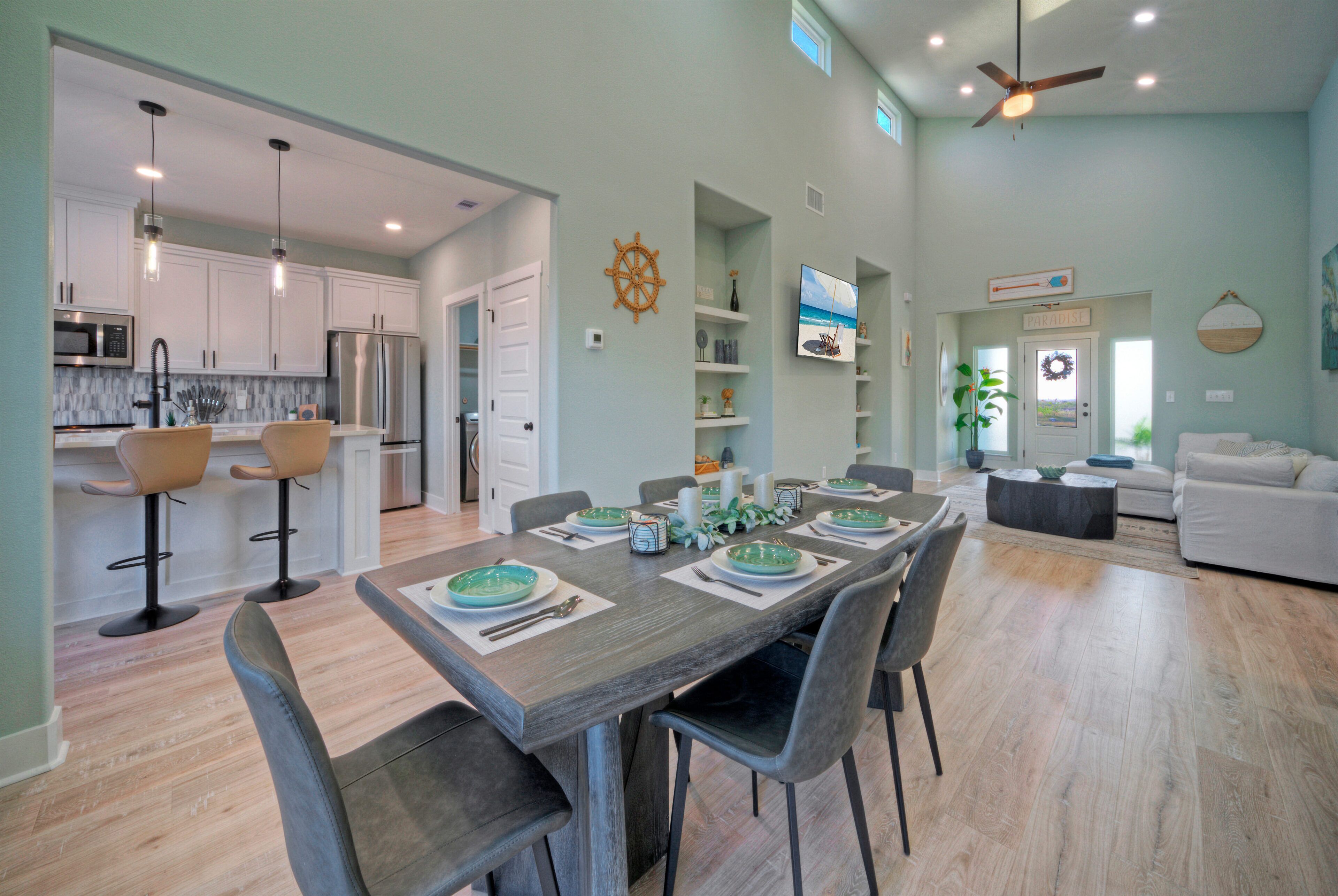 A modern kitchen with stainless steel appliances, a dining area with a wooden table and chairs, and a living room with a white sofa and a ceiling fan.