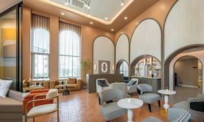 Un vestíbulo de hotel con diseño moderno, que incluye una zona de estar con sofás y sillones, un mostrador de recepción y amplias ventanas que permiten la entrada de luz natural.