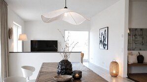 A modern dining area with a wooden table, white chairs, a vase with branches, a candle, and a hanging light fixture. - Vila Karup Modern Scandinavian Holiday Home (Båstad)