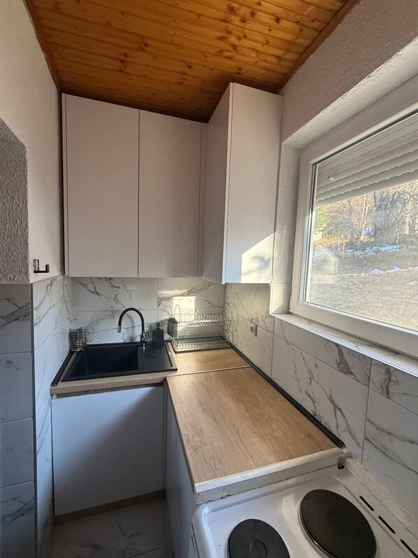 Une cuisine compacte avec des meubles blancs, un plan de travail en bois, un évier et une fenêtre donnant sur les arbres.