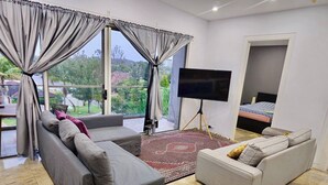 A modern living room with a large TV, two sofas, a glass coffee table, and a view of greenery outside.