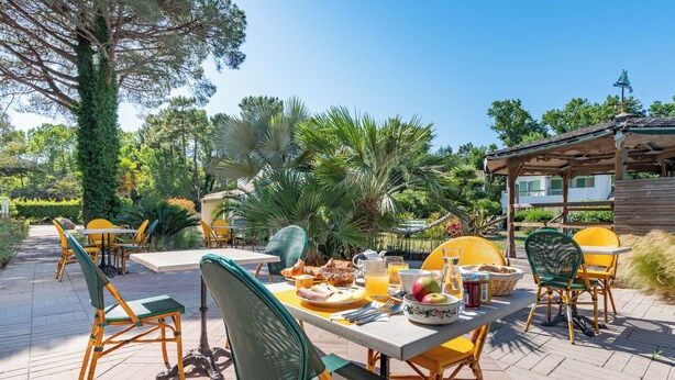 An outdoor dining area with tables and chairs, surrounded by greenery and a building in the background. - Le Parasol (Ars-en-Ré)