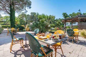 An outdoor dining area with tables and chairs, surrounded by greenery and a building in the background. - Le Parasol (Ars-en-Ré)
