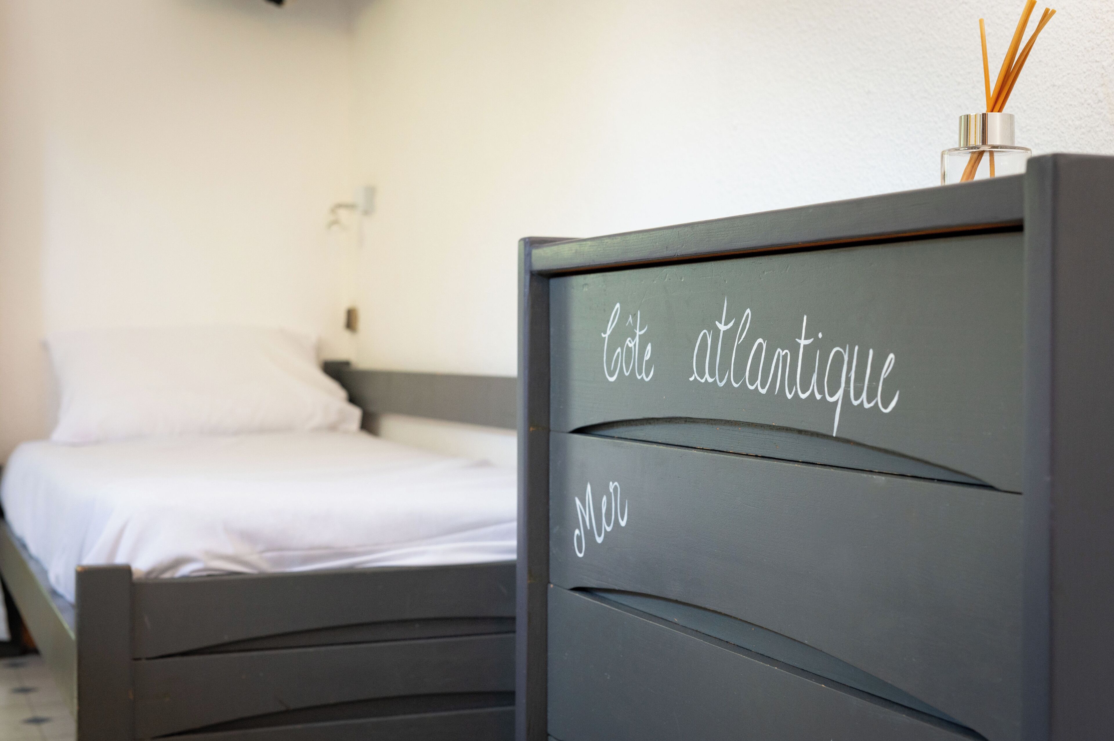 Une chambre à coucher avec un lit, une commode portant une plaque nominative et une lampe fixée au mur.