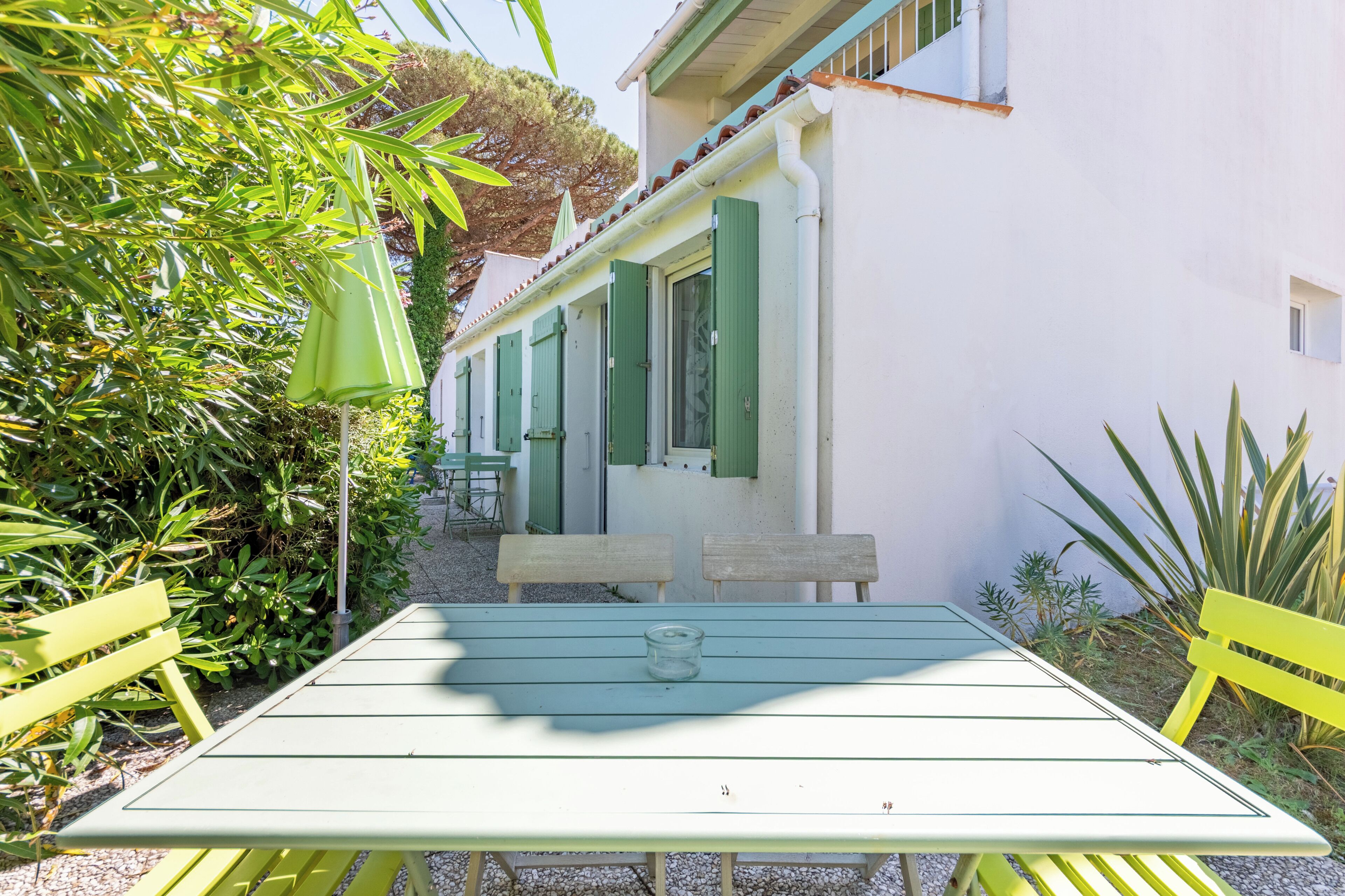 A table with a glass object, green benches, and a building with green shutters.