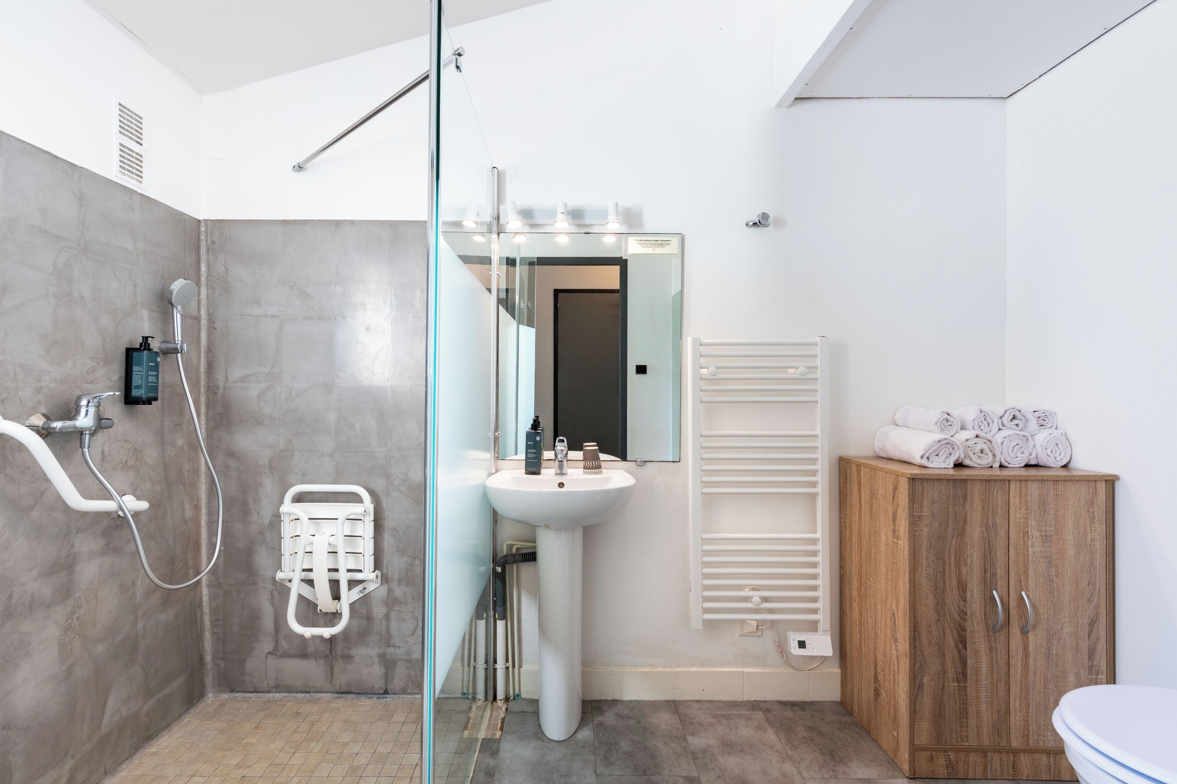 A modern bathroom with a glass shower enclosure, a pedestal sink, a wooden cabinet with towels, and a white radiator.