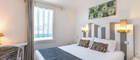 A hotel room with a bed, bedside tables, a chair, a window with curtains, and a wall-mounted artwork.