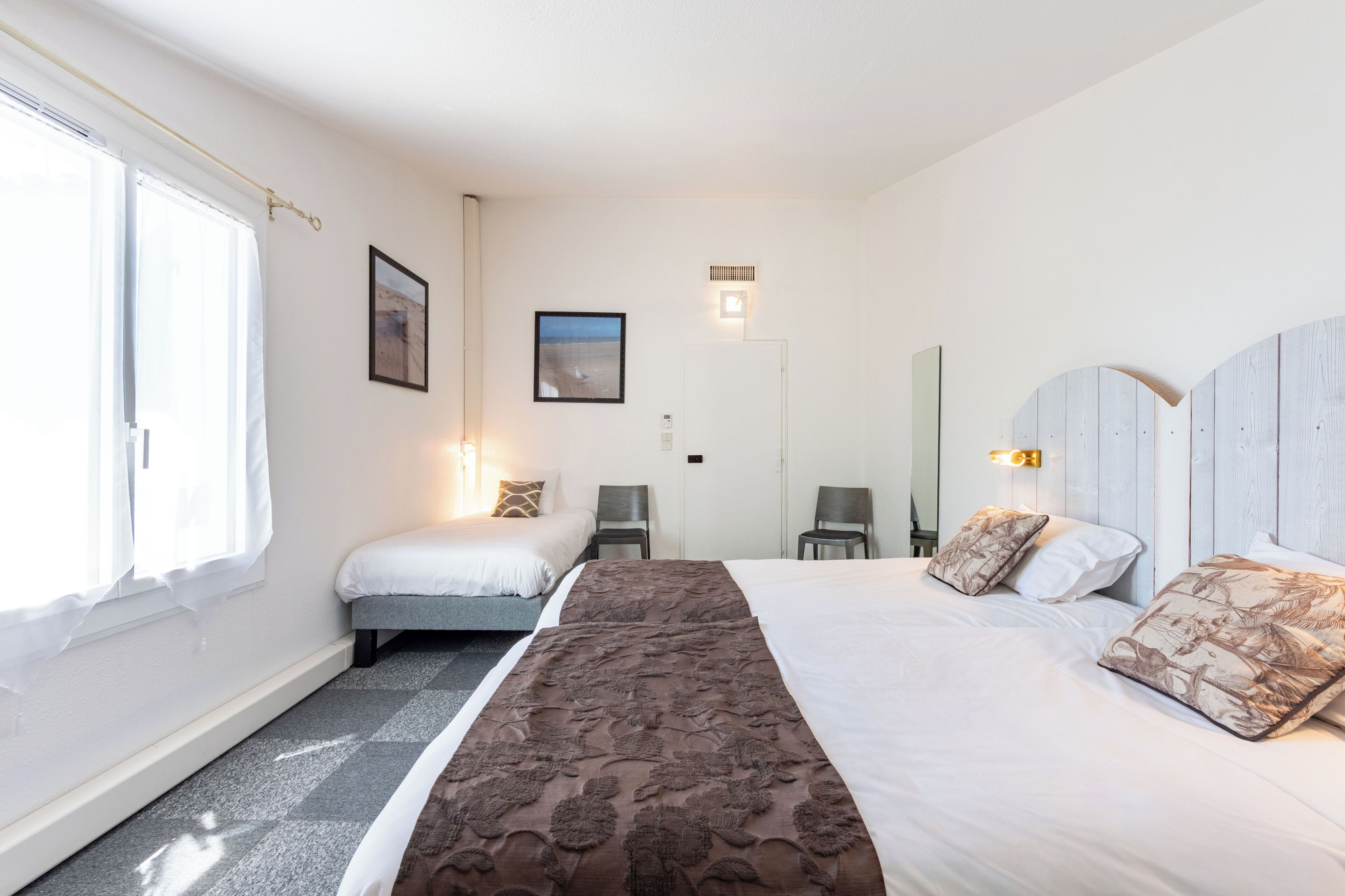 Ein Hotelzimmer mit einem großen Bett, zwei Einzelbetten, einem Fenster mit Vorhängen, einem Wandbild und einem Spiegel.