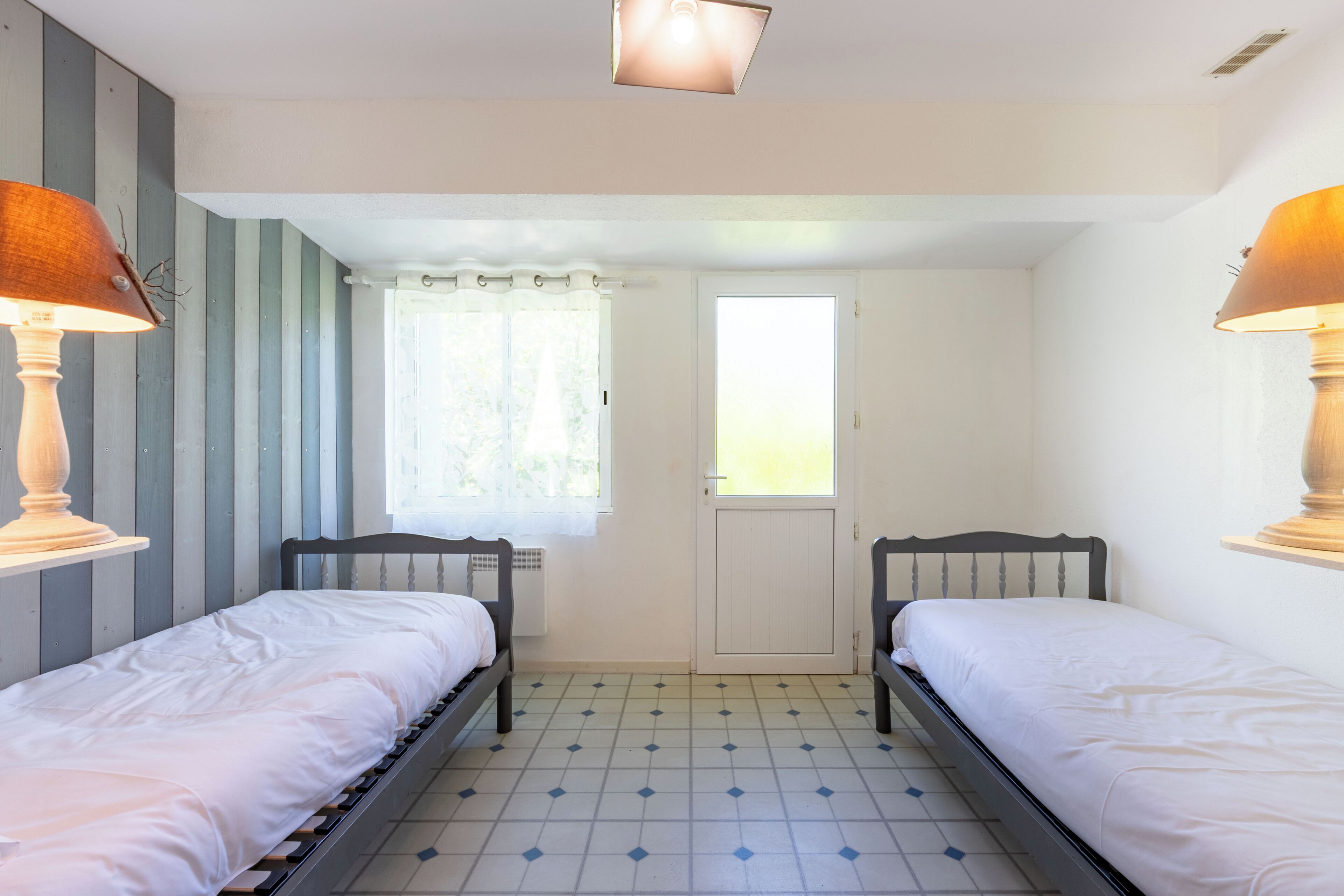 A room with two single beds, a window with curtains, a door, and a light fixture on the ceiling.