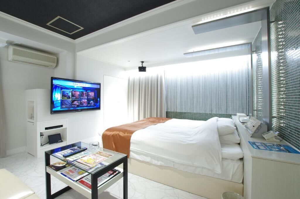 A hotel room with a bed, a TV mounted on the wall, a bedside table with a magazine, and a window with curtains.