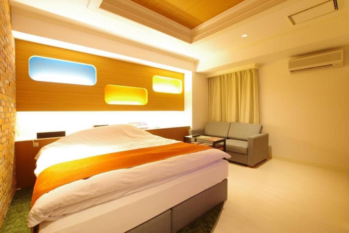 A hotel room with a bed, a sofa, a small table, and a wall-mounted air conditioner.