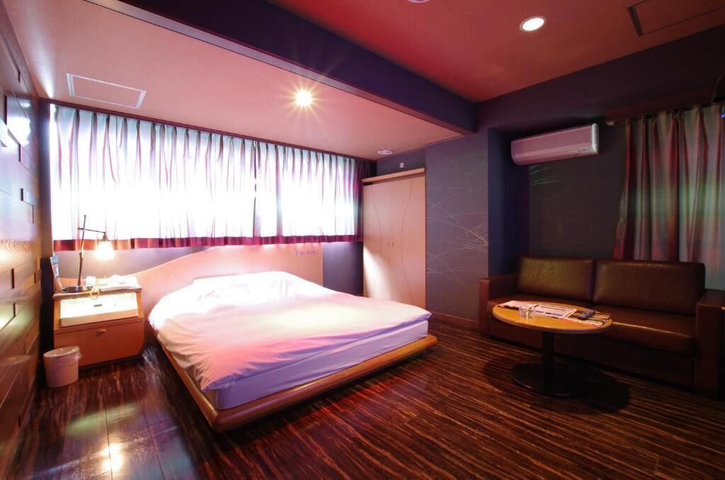 A hotel room with a bed, a nightstand, a sofa, and an air conditioning unit.