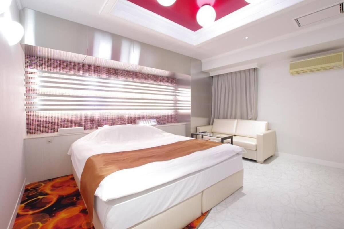 A modern hotel room with a large bed, a sofa, a ceiling light fixture, and a window with blinds.