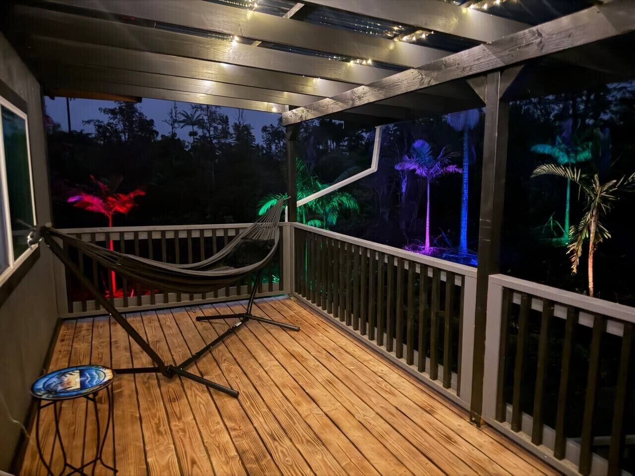 A wooden deck with a hammock and a small table, surrounded by a railing and string lights.