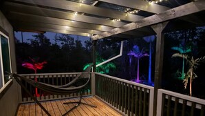 A wooden deck with a hammock and a small table, surrounded by a railing and string lights. - Jungle Lux Cabin - Private Retreat w/ AC & WiFi (Keaau)