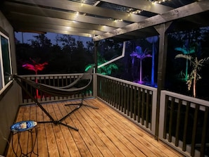 Una terraza de madera con una hamaca y una mesita, rodeada por una barandilla y luces colgantes.