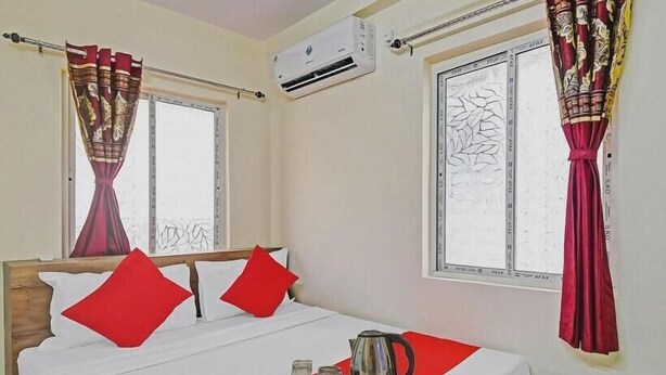 A hotel room with a bed, red pillows, a tray with a kettle, cups, and a tray, an air conditioner, and a window with curtains. - sapphire guest house (Kolkata)