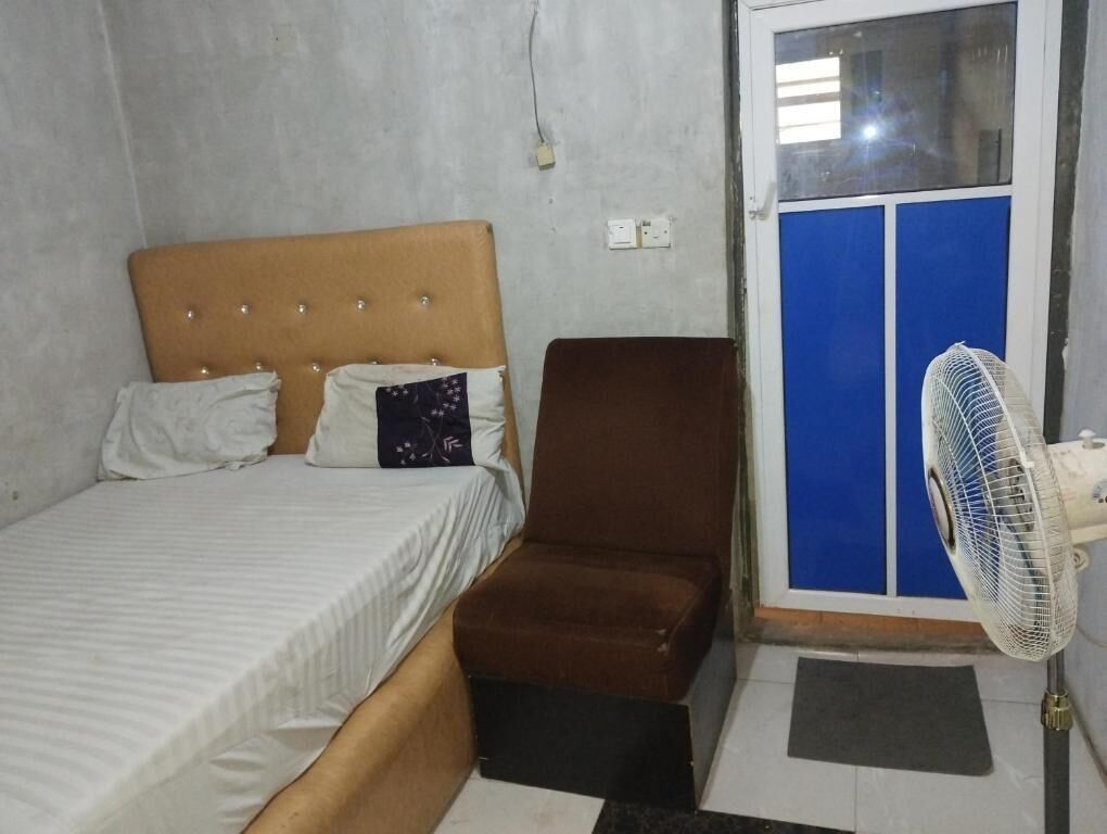 Une chambre à coucher avec un lit, un fauteuil marron, un ventilateur sur pied et une porte bleue.