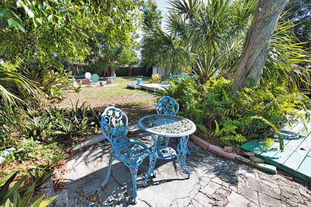 Een terras met een stenen vloer, twee blauwe metalen stoelen, een ronde tafel en omliggend groen.