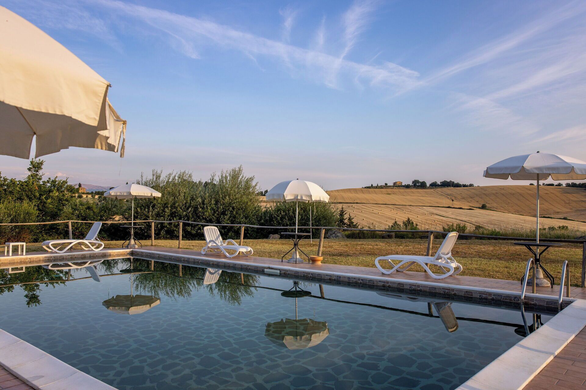 An outdoor infinity pool with lounge chairs and umbrellas, surrounded by a landscaped garden and a view of rolling hills.