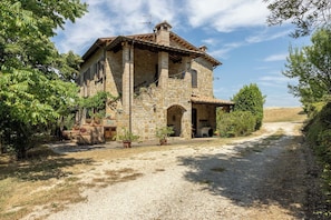 Un bâtiment en pierre doté d’un porche couvert, d’une allée en gravier et entouré de verdure.
