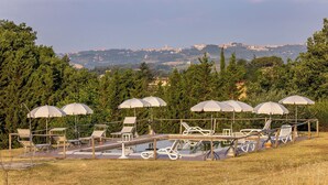 Ein Poolbereich mit Liegestühlen und Sonnenschirmen, umgeben von viel Grün und Blick auf ferne Hügel.