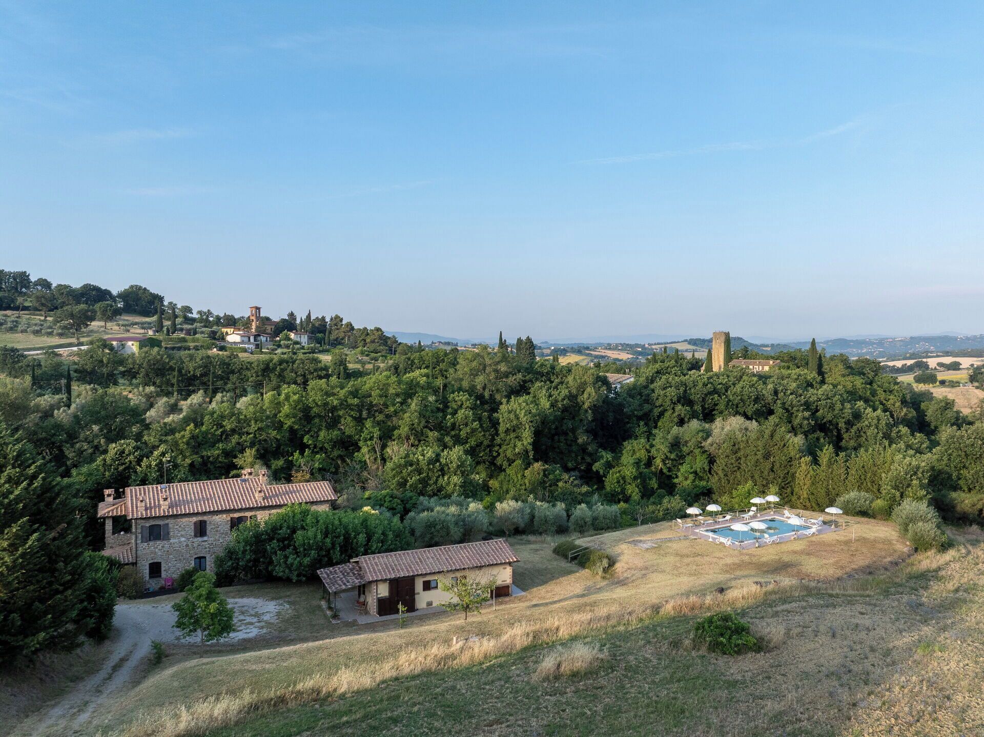 A rural landscape with a stone house, a swimming pool with umbrellas, and a distant tower.