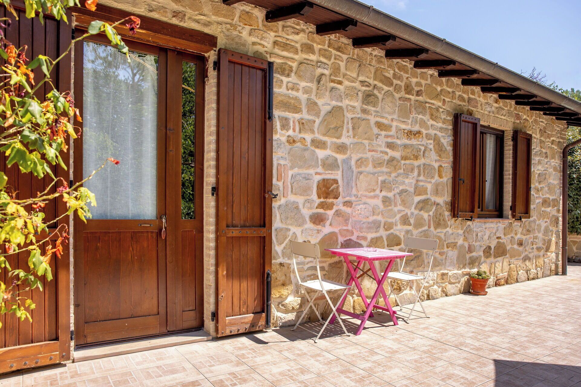 Un bâtiment en pierre avec des portes et des fenêtres en bois, une terrasse carrelée et une table rose avec des chaises.