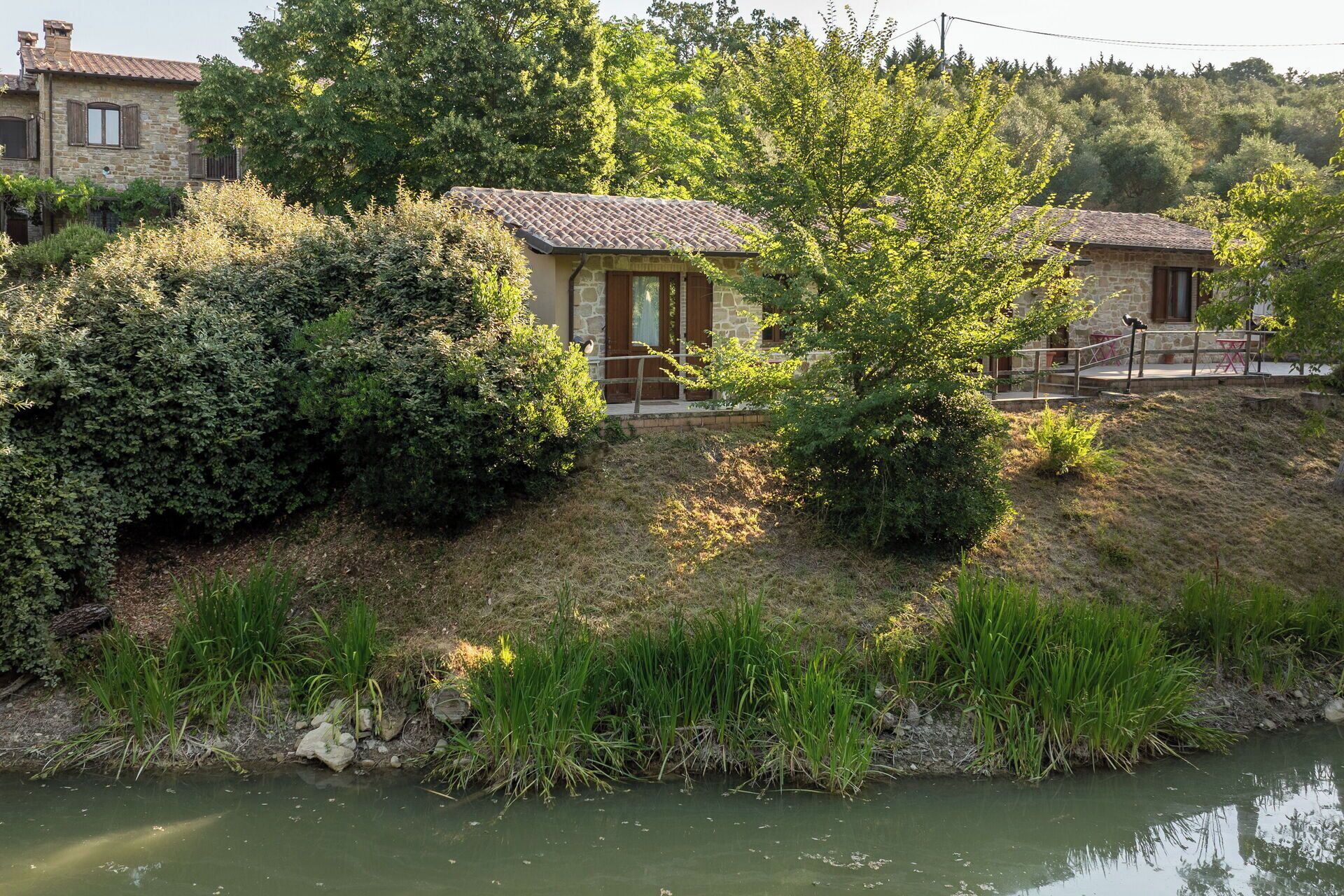 A stone building with a tiled roof, situated beside a river, surrounded by dense greenery.