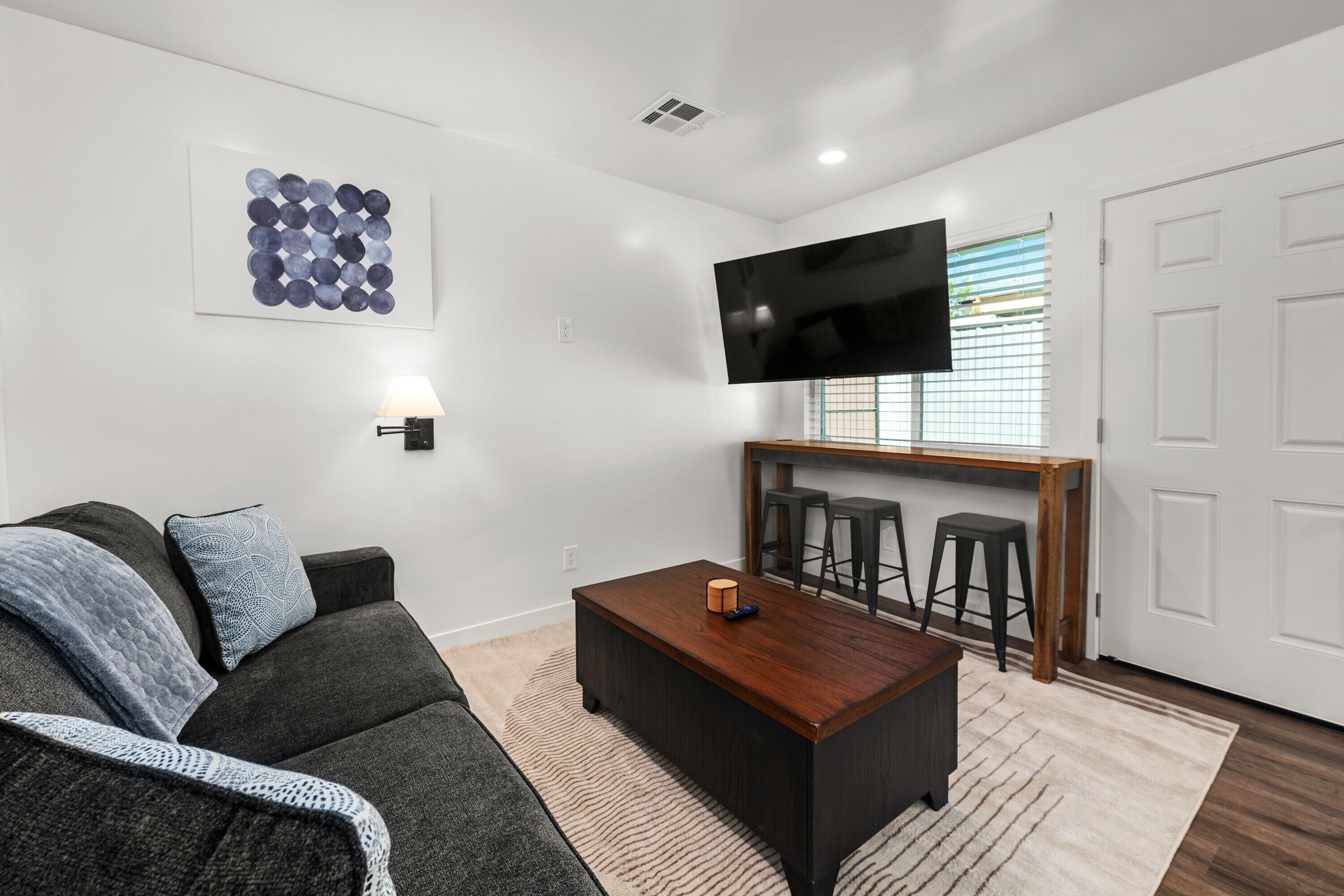 A modern living room with a dark sofa, a coffee table, a TV mounted on the wall, and a bar area with stools.