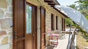 Ein Steinhaus mit Holztüren und -fenstern, ein überdachter Außenbereich mit Tisch und Stühlen sowie ein weißer Sonnenschirm, der Schatten spendet.