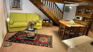 Una sala de estar acogedora con un sofá verde, una mesa de comedor con sillas, una escalera y una zona de cocina con microondas.