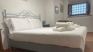 Una cama con sábanas blancas, cabecera de metal azul y una mesita de noche pequeña, junto a una ventana con diseño de celosía.