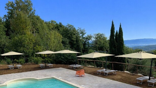 Una piscina rectangular con agua azul cristalina, rodeada de sillas de descanso y sombrillas, con árboles y un cielo despejado al fondo.