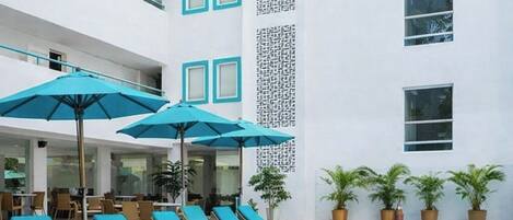 A poolside area with lounge chairs and umbrellas, a swimming pool, and a building with a white facade and patterned wall.