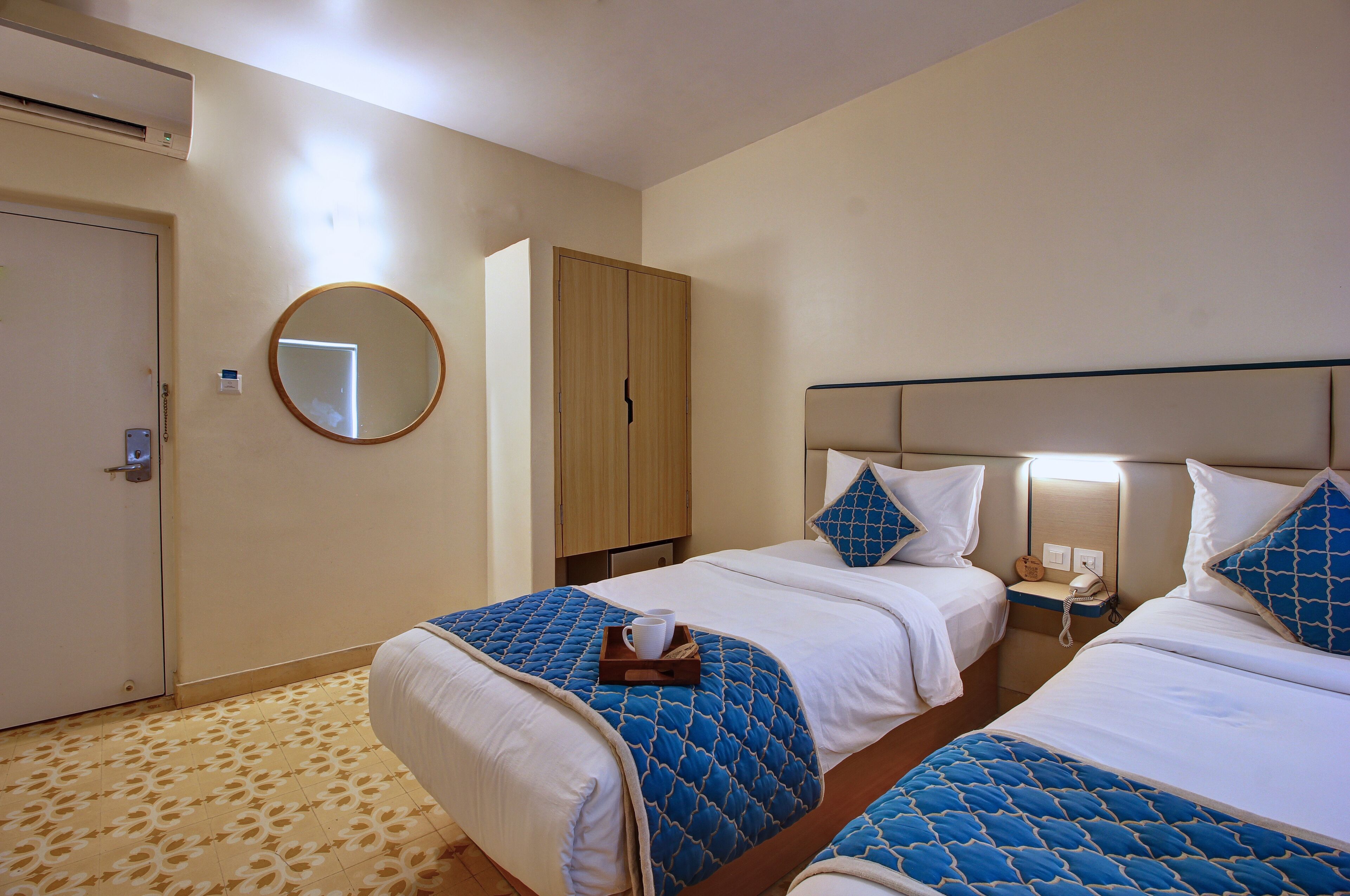 A hotel room with two beds, a wardrobe, a round mirror, and a bedside table with a lamp.