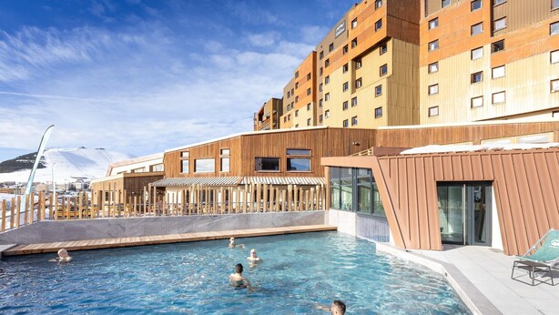 A swimming pool with people, a wooden deck, and a modern building with multiple floors.