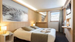 A hotel room with a bed, bedside lamps, a mounted TV, a window with curtains, and a large framed picture on the wall.