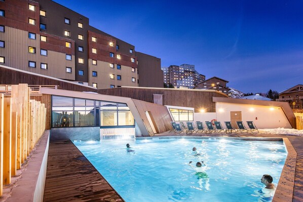 A swimming pool with people, wooden deck, and modern buildings in the background.