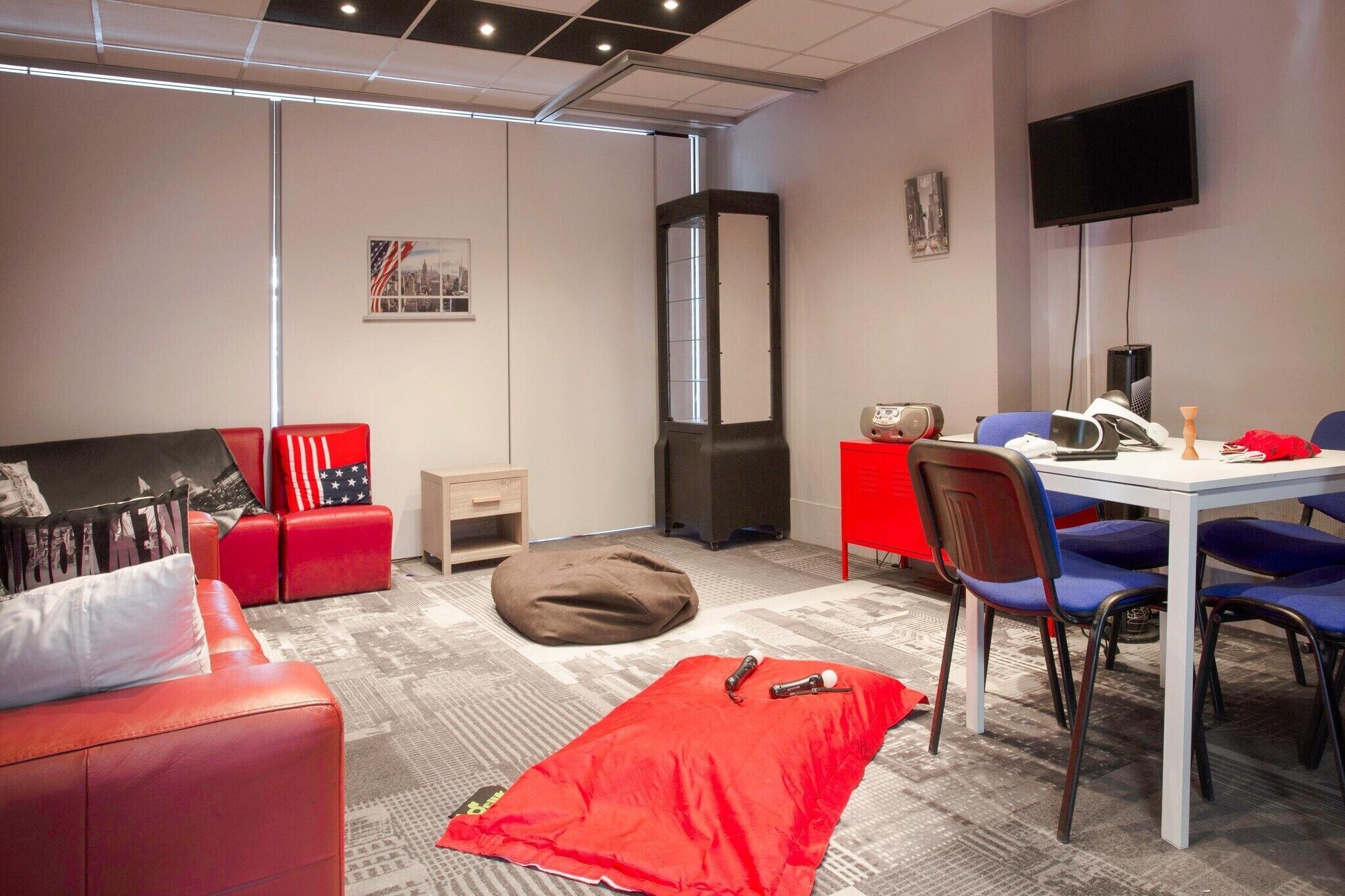 Une pièce moderne avec un canapé rouge, un fauteuil poire, une table blanche avec des chaises bleues, un téléviseur et un meuble rouge.