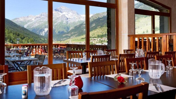 A restaurant with tables and chairs, large windows with a mountain view, and a wooden deck. - Alpine Base with Hot Tub & Pool (LANSLEBOURG)