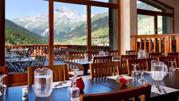 En restaurant med borde og stole, store vinduer med udsigt til et bjerglandskab, og en træterrasse.
