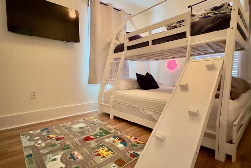 Une chambre avec un lit superposé, un téléviseur fixé au mur, une fenêtre avec des rideaux et un tapis de jeu sur le sol.