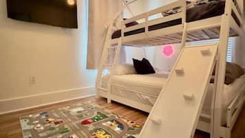 Une chambre avec un lit superposé, un téléviseur fixé au mur, une fenêtre avec des rideaux et un tapis de jeu sur le sol.