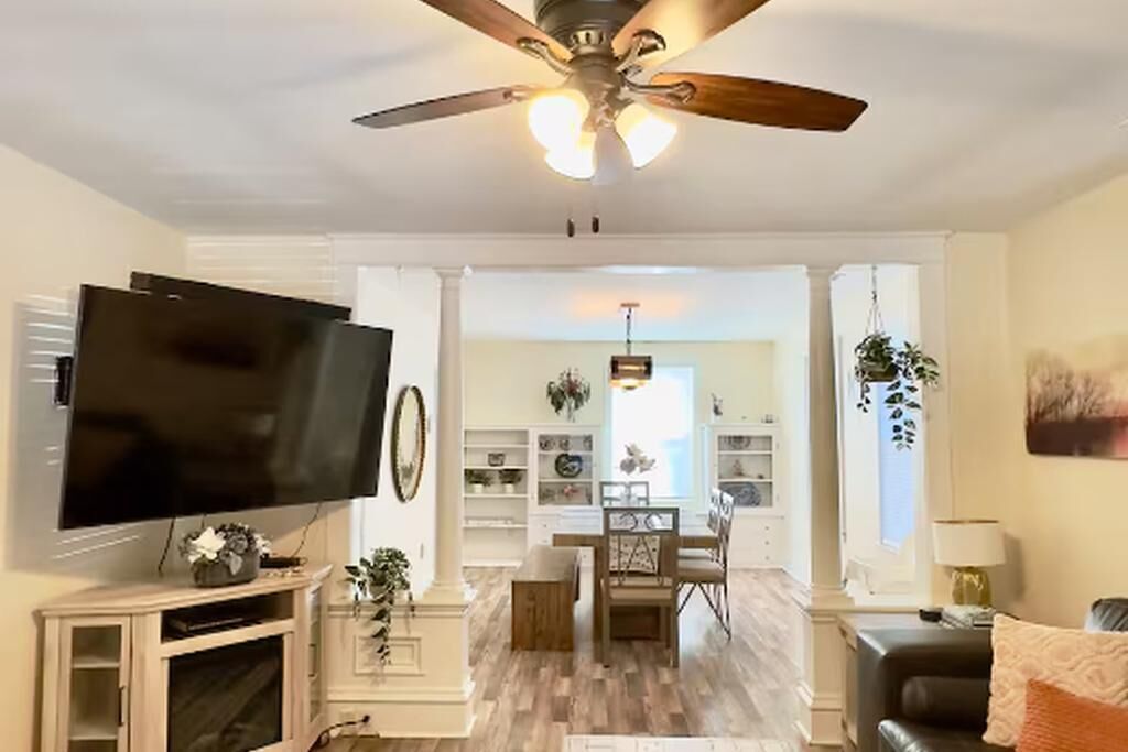 Un salon équipé d’un téléviseur à écran plat, d’un ventilateur de plafond, d’un plancher en bois et d’un coin repas avec une table et des chaises.