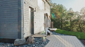 Ein modernes Haus mit Holzfassade, einem gepflasterten Eingangsbereich und einem Garten mit Steinen und Bäumen.