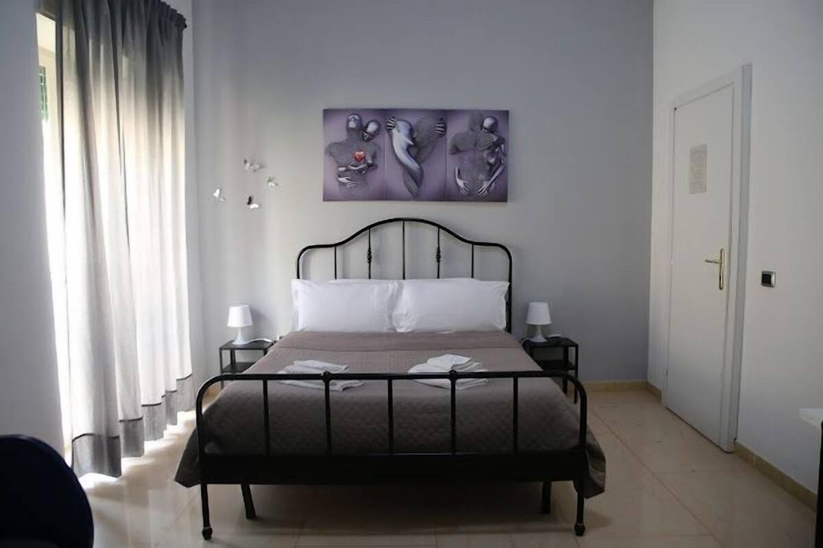 A bedroom with a bed, two bedside tables, a wall-mounted artwork, a door, and a window with curtains.